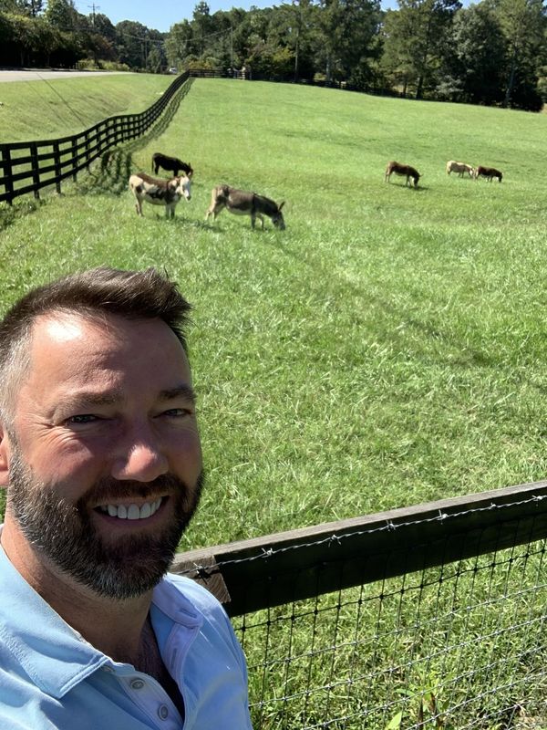 Author Matt Clement smiling at the meadow fence, with the miniature donkeys grazing behind him